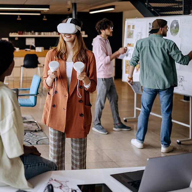 Menschen stehen an einem Flipchart und eine Person testet eine VR-Brille